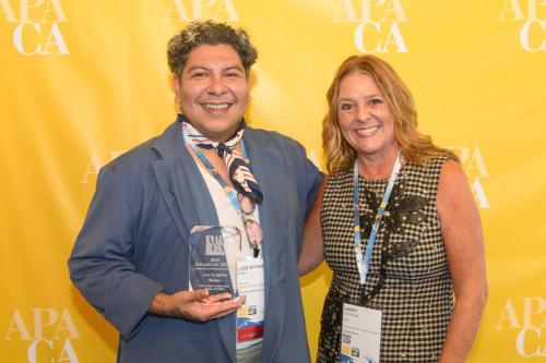 Two smiling people stand in front of a yellow backdrop, one holding an award.