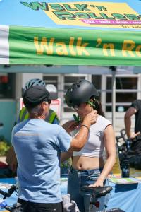 Man helps woman adjust bike helmet under green event tent.