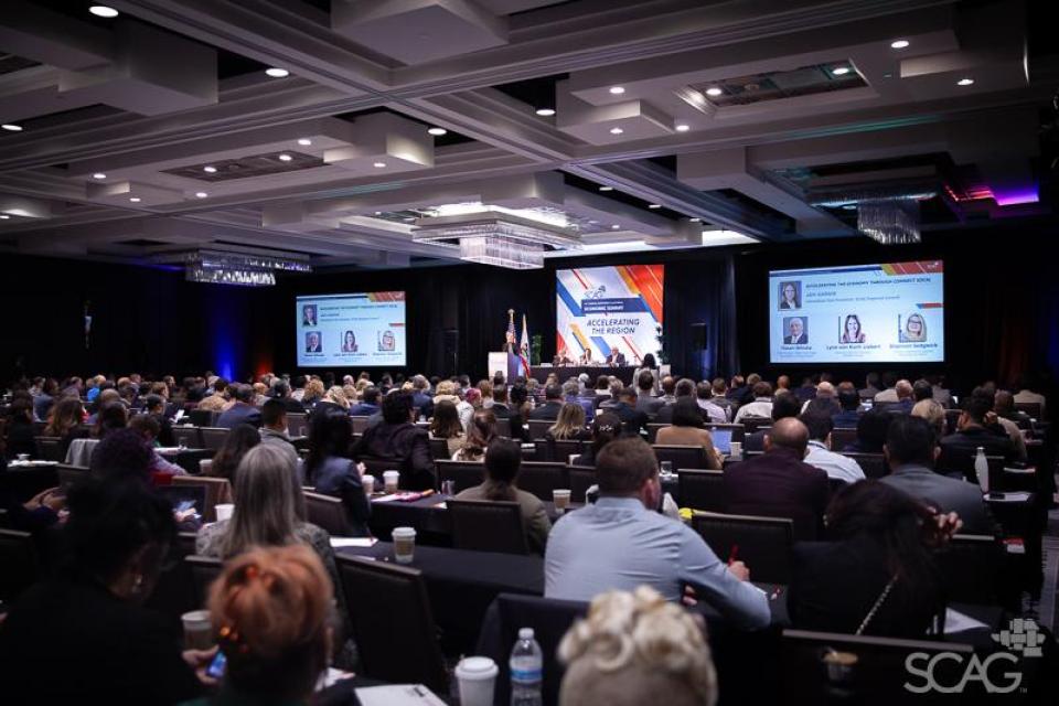 Overall room  at the Economic Summit filled with attendees, a stage and two screens.